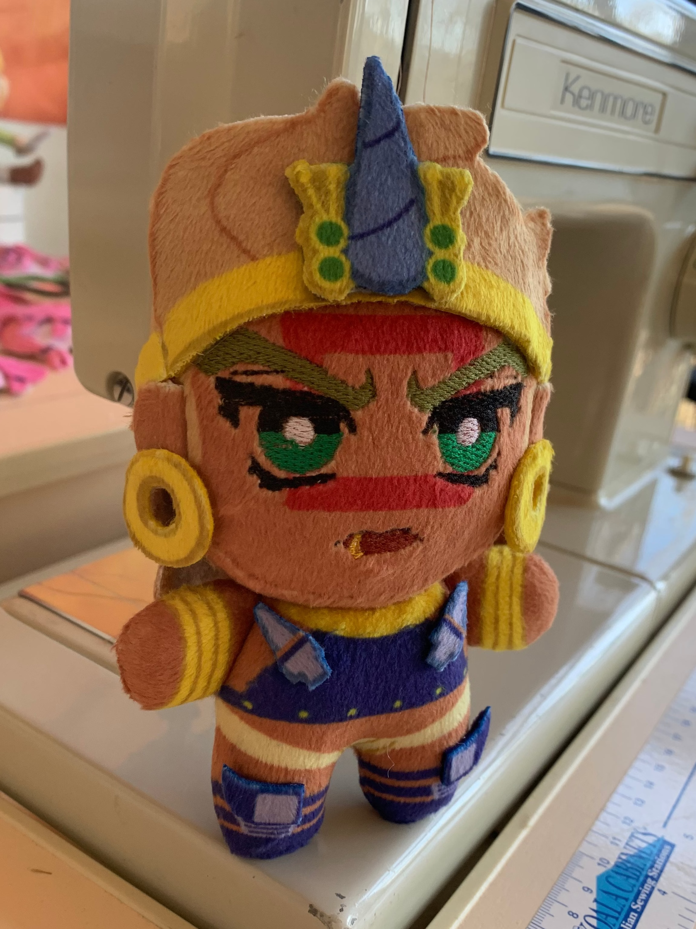 A plush toy with golden jewelry and a red square face marking standing in front of a sewing machine