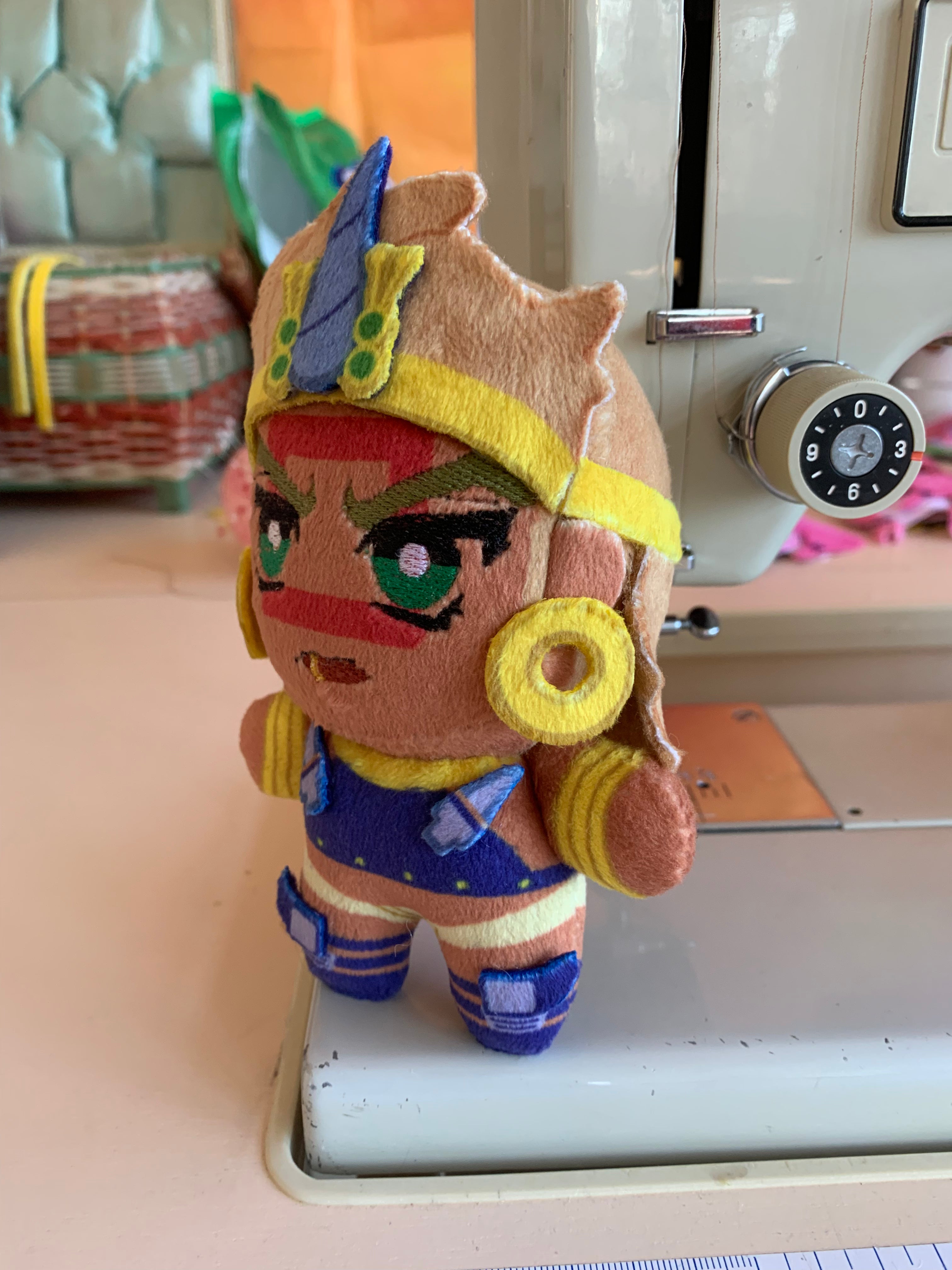 Side view of a plush toy with golden jewelry and a red square face marking standing in front of a sewing machine