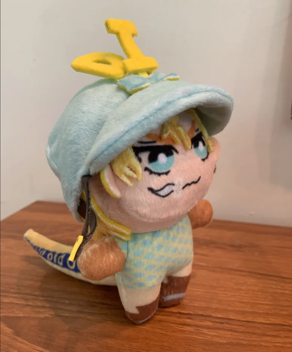 Plush toy with a blue jockey's cap and yellow dinosaur tail standing up on a wooden surface