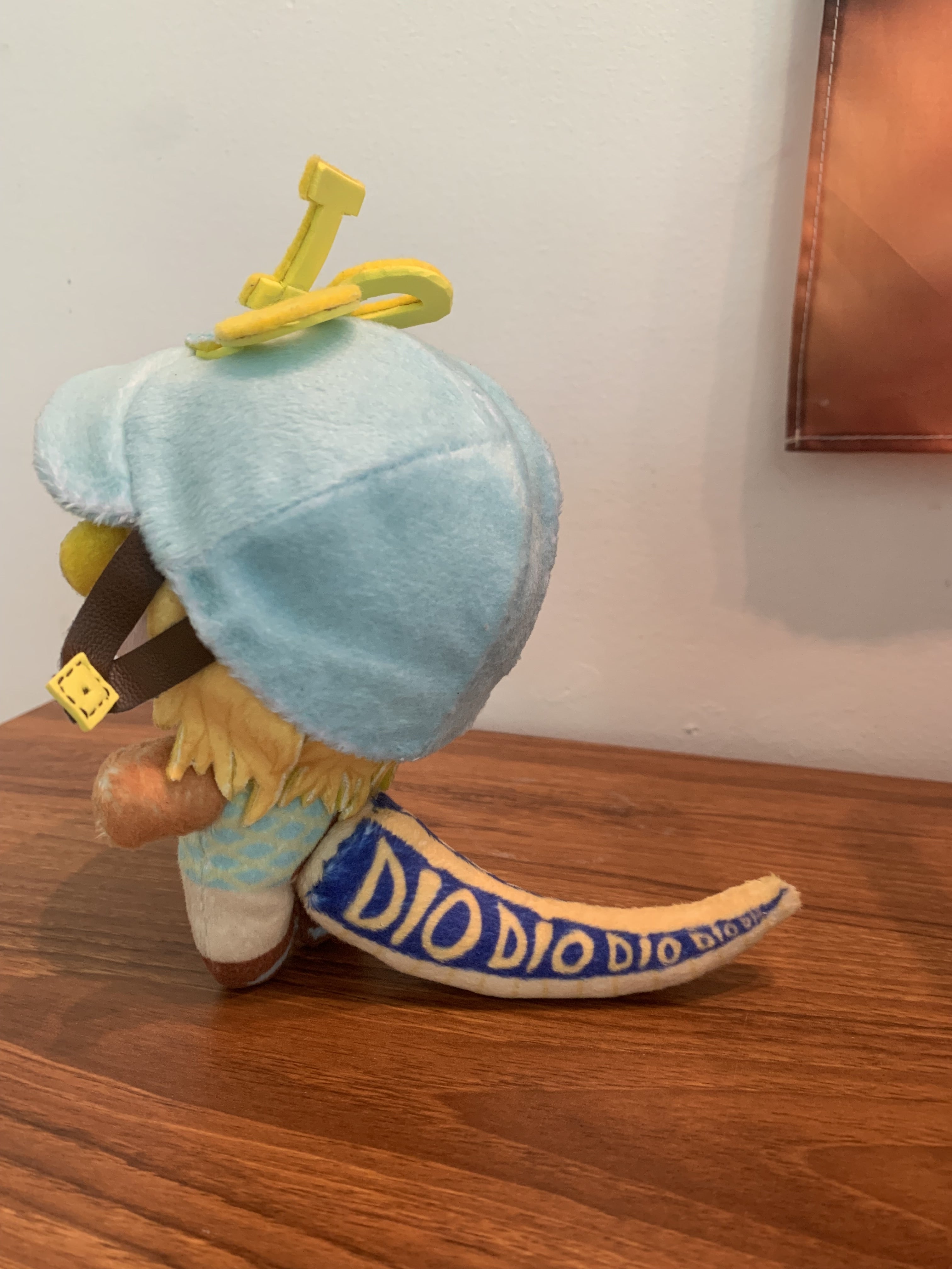 Side view of a plush toy with a blue jockey's cap and yellow dinosaur tail standing up on a wooden surface