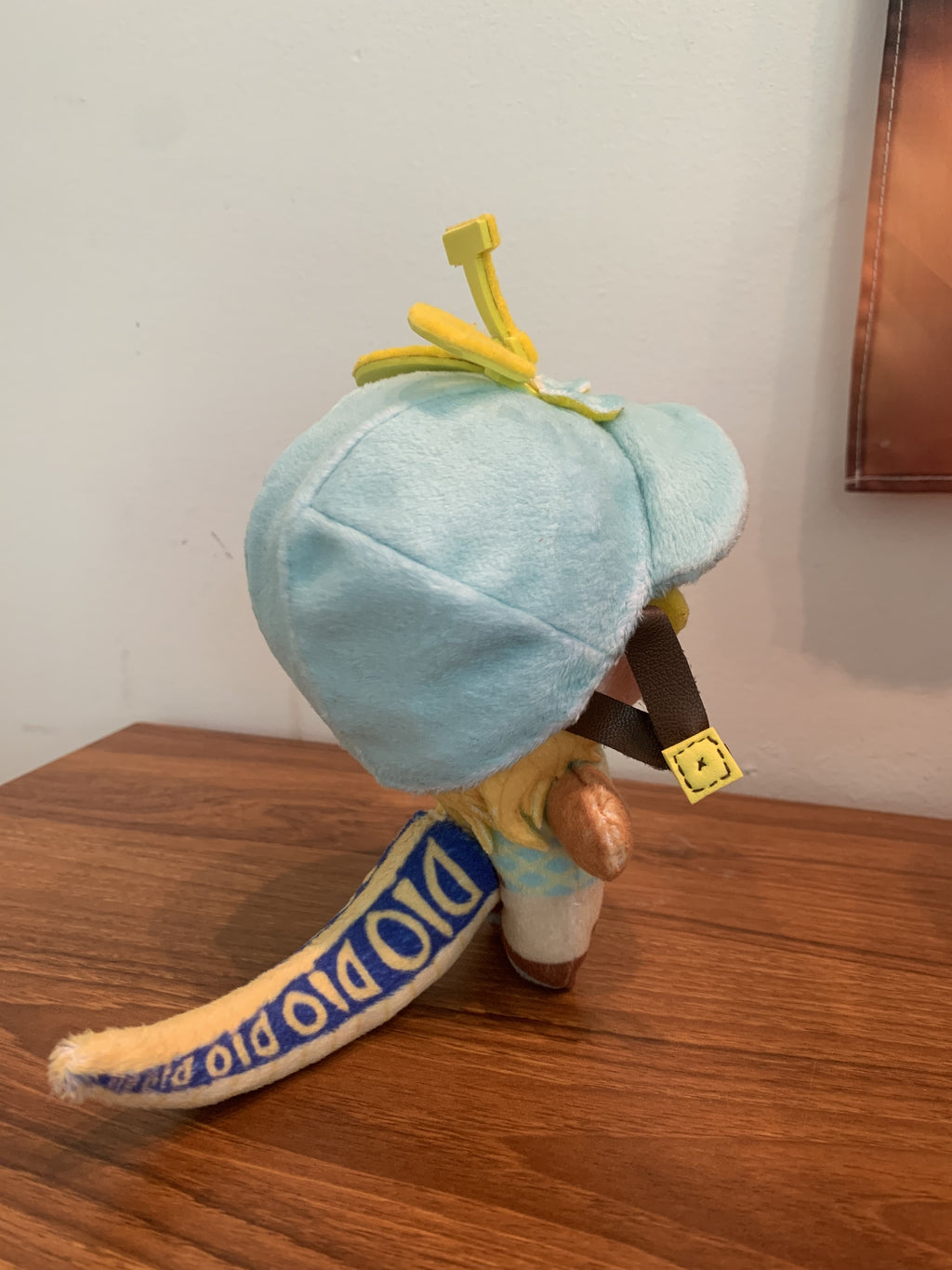 Side view of a plush toy with a blue jockey's cap and yellow dinosaur tail standing up on a wooden surface